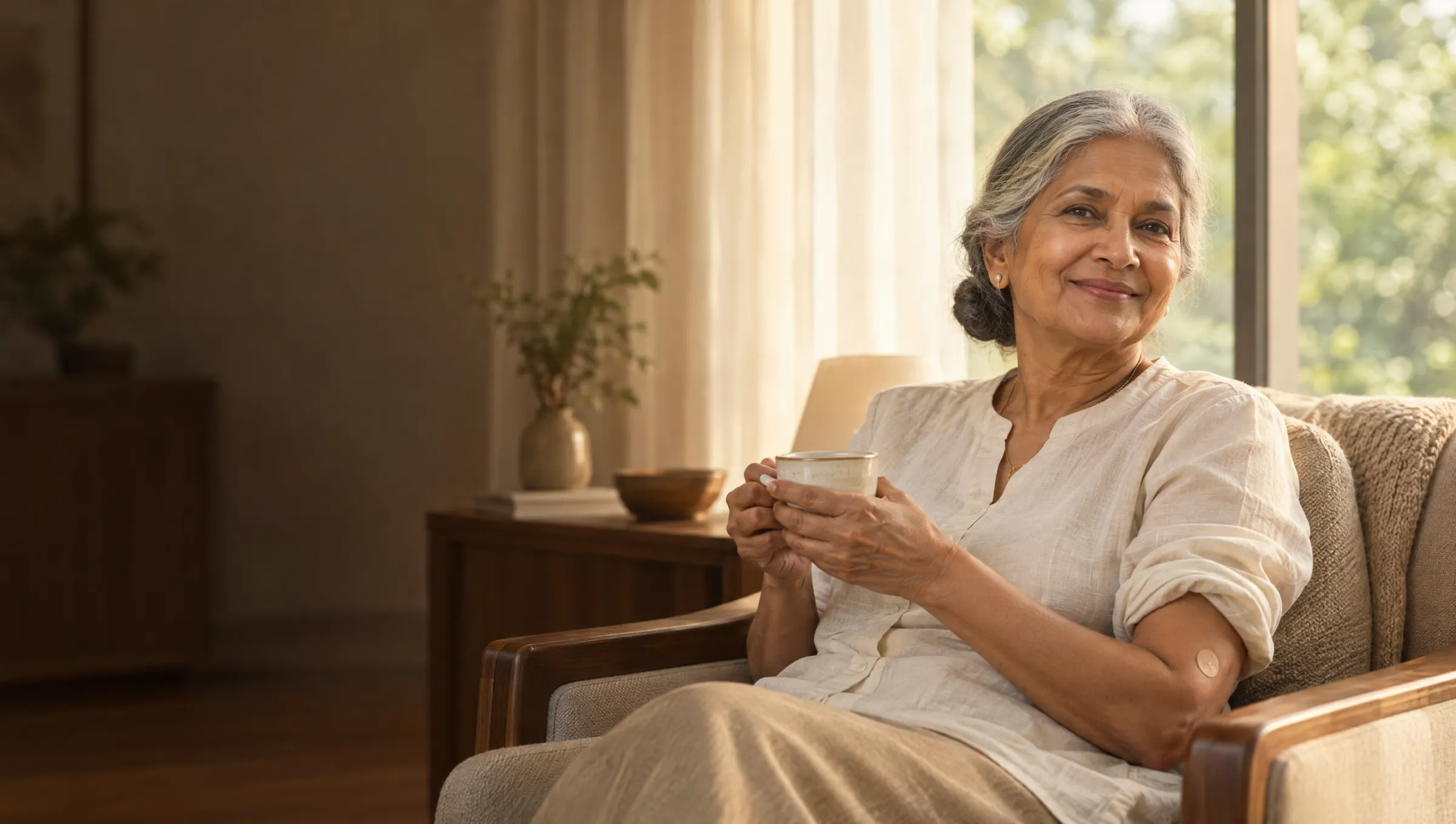 An older woman smiling by a window with a cup of tea, a small plaster on her arm after a blood test