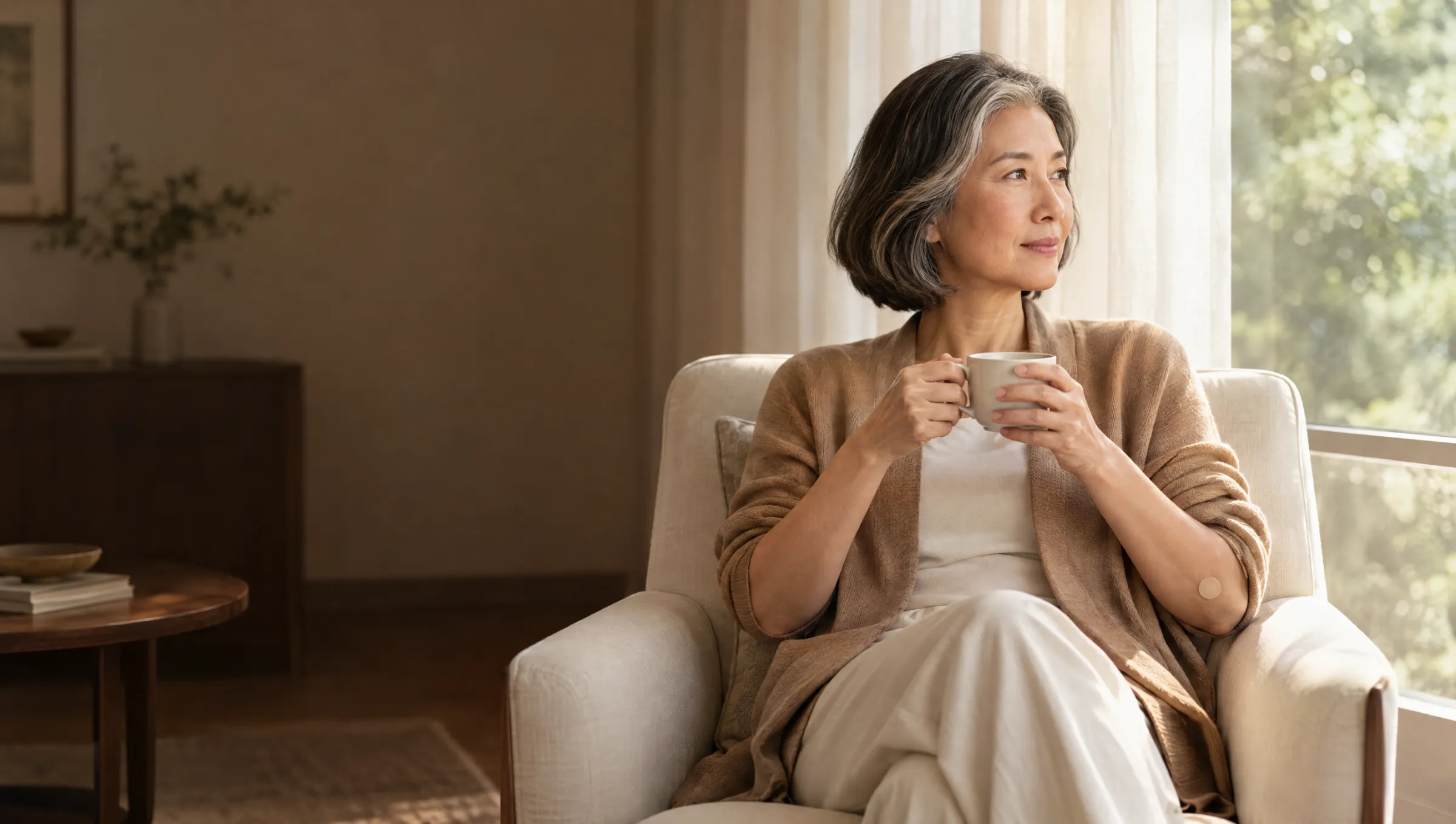 A woman in her mid-fifties resting by a window with a cup of tea, a small plaster on her arm