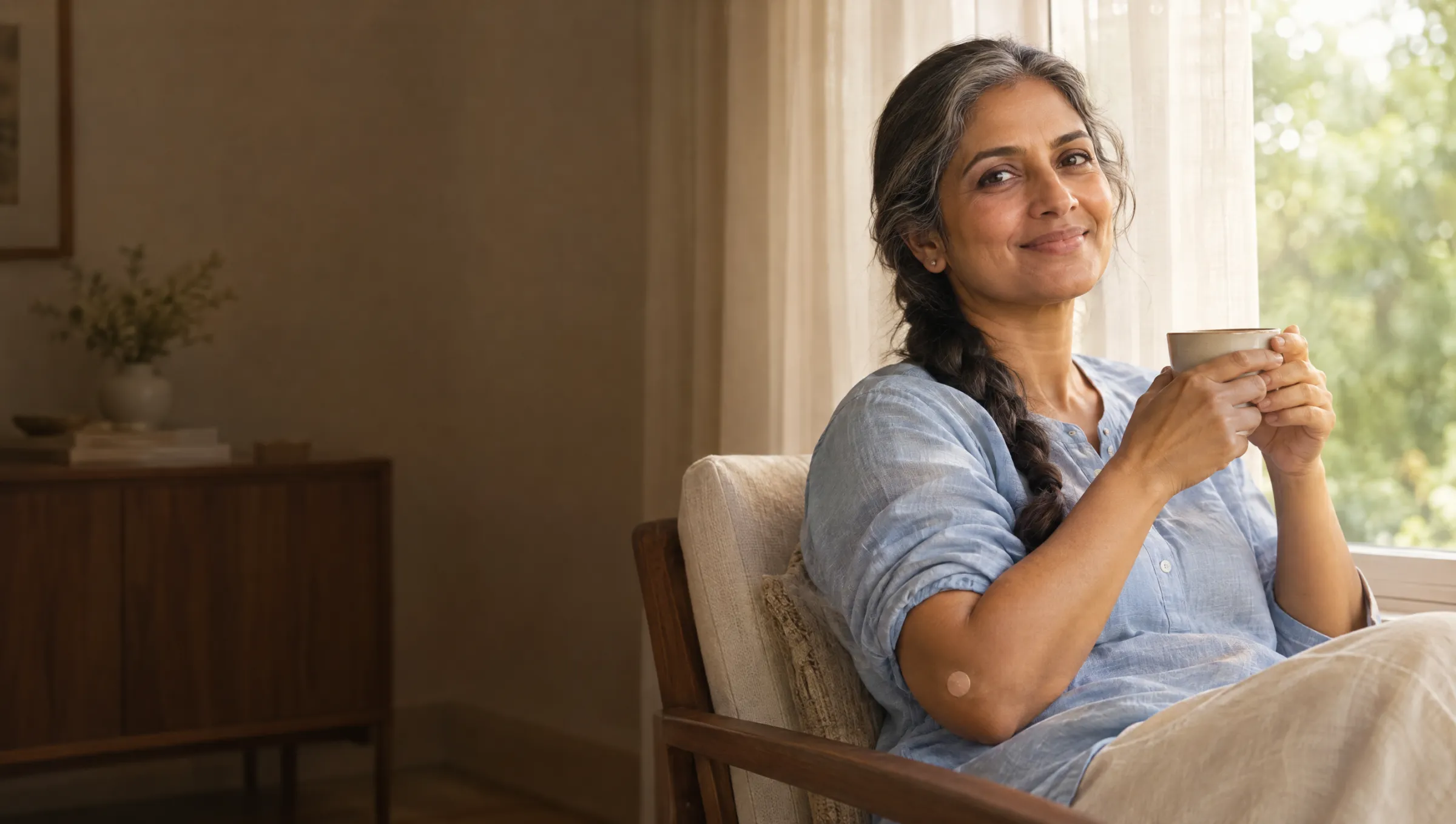 A woman in her fifties with a cup of tea in a bright lounge, a small plaster on her arm after a blood test
